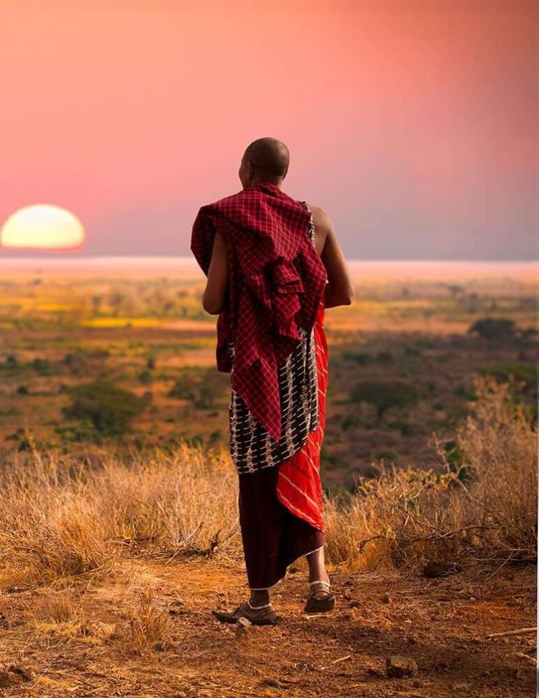 Maasai man in a red shuka walking towards the sunrise across the plains